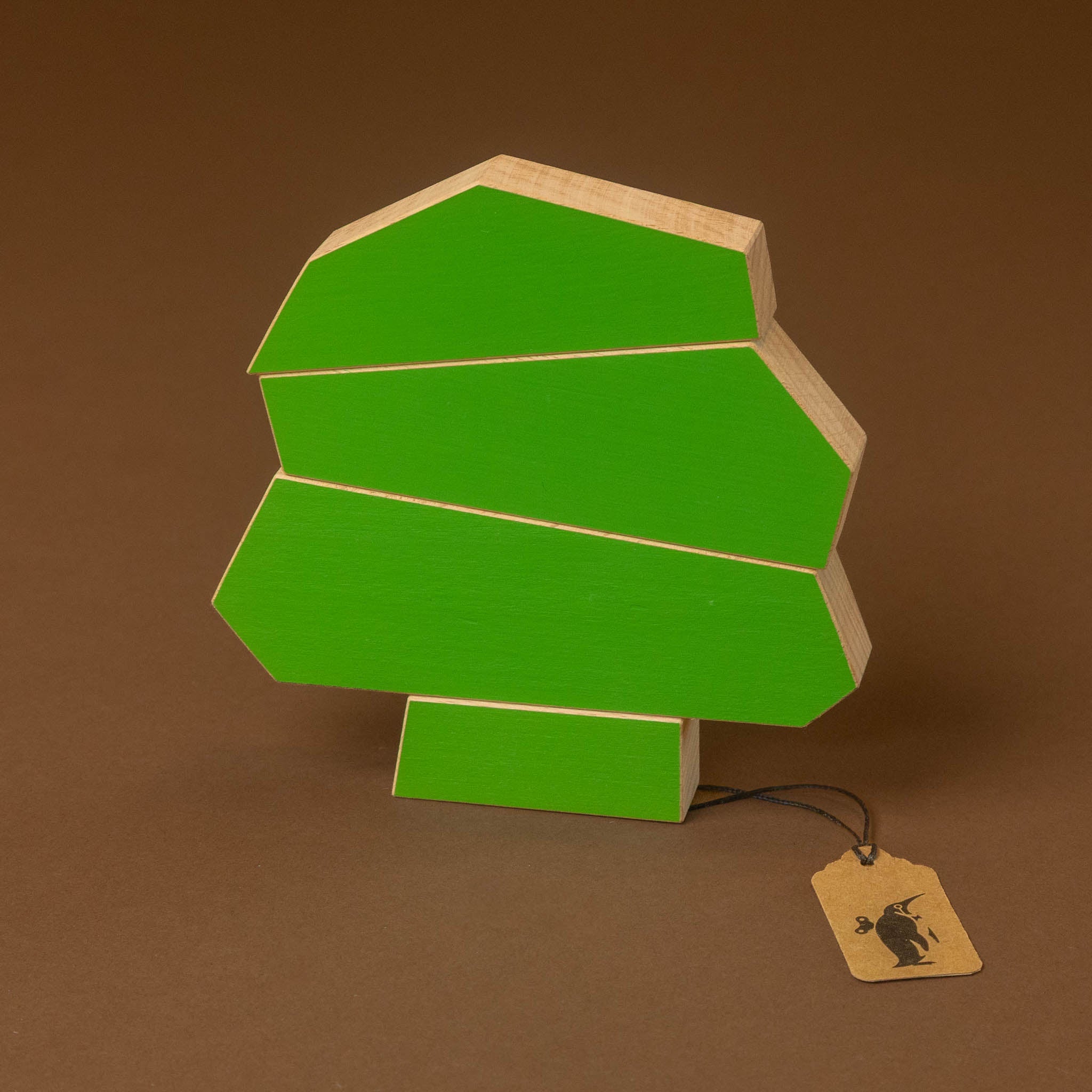 A green geometric tree-shaped object with light natural wood edges, standing on a small rectangular base against a solid brown background. A small paper tag with a black rabbit logo hangs from a string.