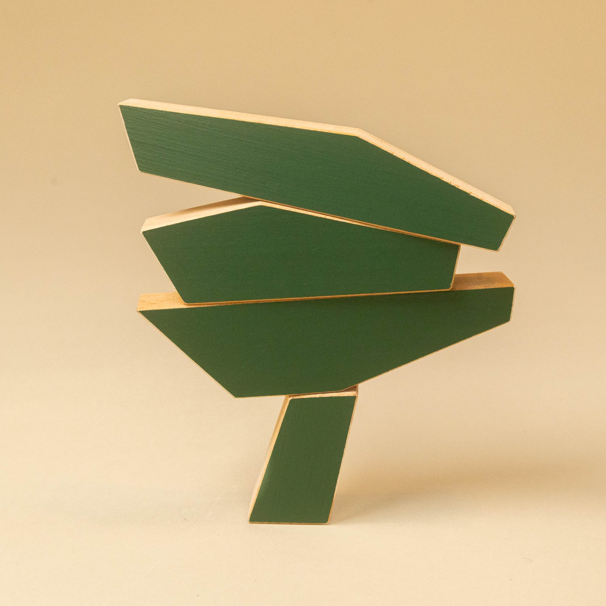 A modern abstract geometric sculpture made of layered green wooden planks with natural light wood edges, set against a solid warm beige background.