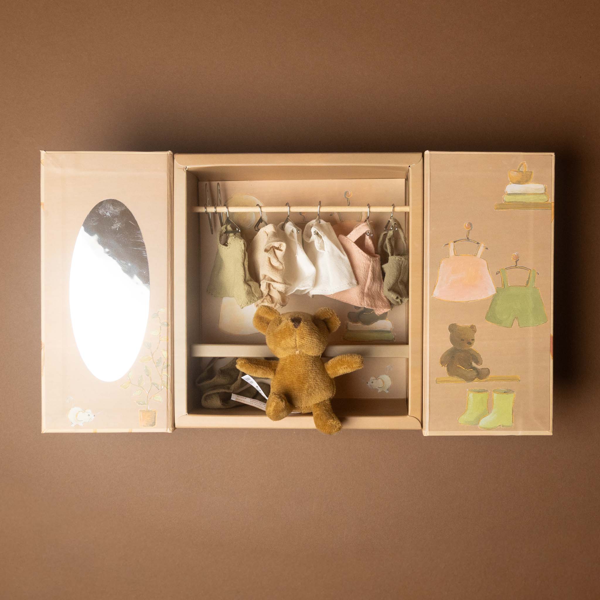 A miniature toy woodland-bears-wardrobe in a light wood tone, featuring a hanging rod with baby clothes, a plush teddy bear, and illustrated clothing on the side panels, shown against a solid brown background.