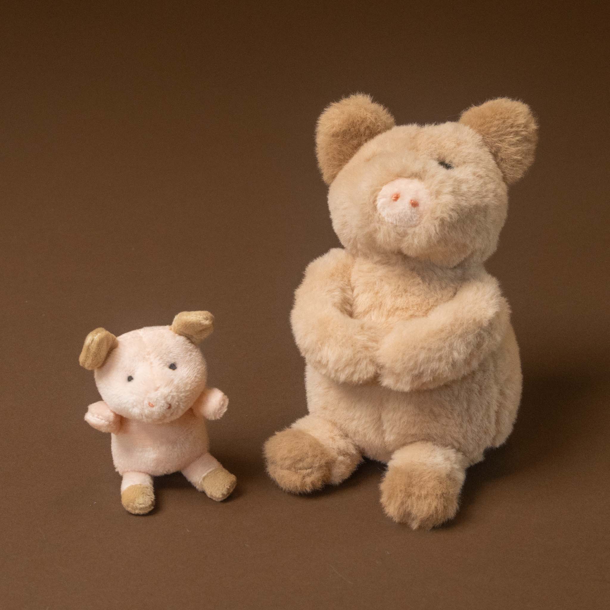 woodland-friends-pig-baby on a solid brown studio background, with a larger cream-colored pig sitting on the right and a smaller pale pink pig standing on the left.