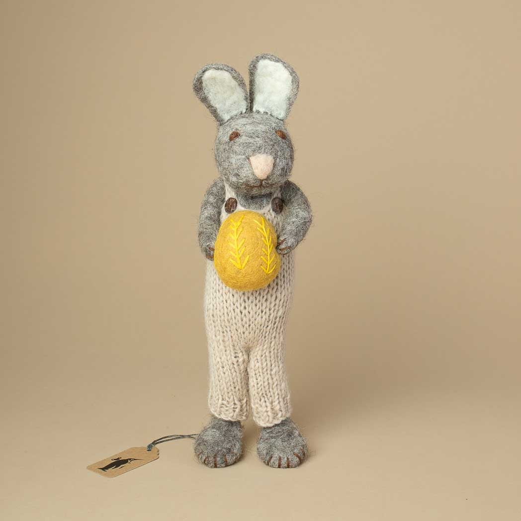 grey-felted-bunny-with-long-ears-wearing-a-knitted-overall-and-holding-a-yellow-embroidered-easter-egg-in-his-hands